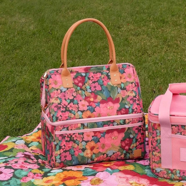 A pink floral insulated tote bag with tan handles sits on a grassy lawn next to a matching pink floral lunch bag.