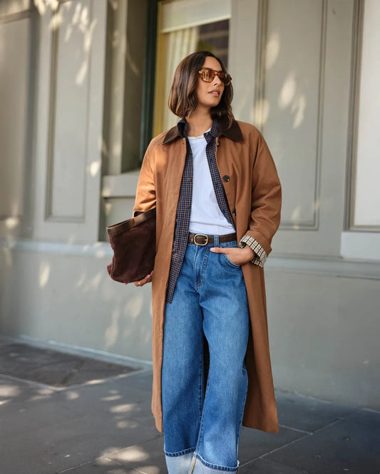 A woman stands on a city sidewalk wearing a long tan coat over a white top, blue plaid shirt, and blue jeans, holding a brown bag. The background shows a neutral-toned building facade with soft shadows.