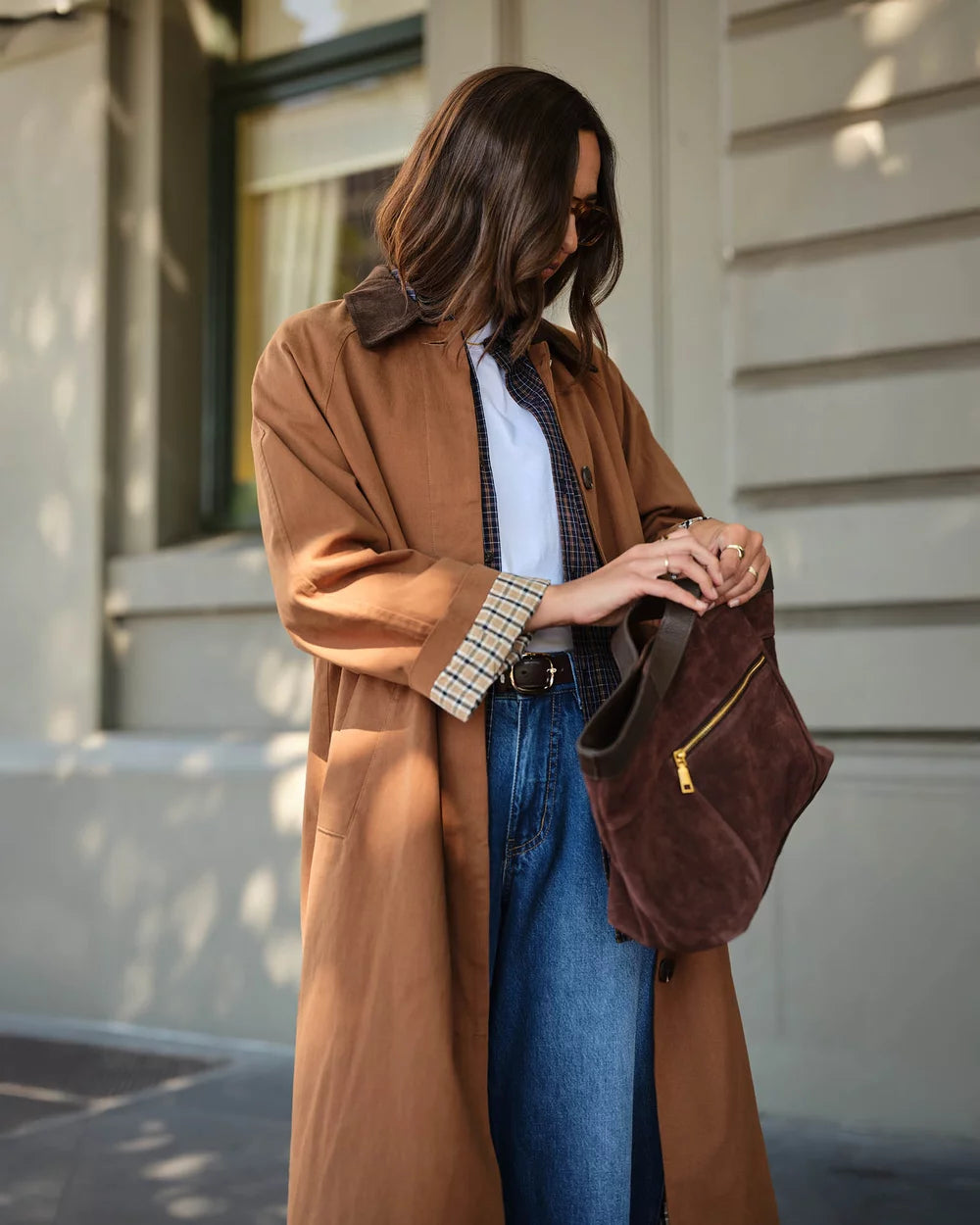 A woman wearing a camel brown trench coat, blue jeans, and a patterned shirt holds a dark brown suede shoulder bag with a gold zipper, set against a modern building facade.