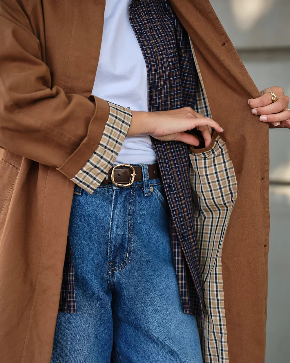 A close-up of a person wearing a brown trench coat over a white top and blue jeans, with a plaid shirt tucked inside the coat. The person is shown from the chest down, with one hand opening the coat to reveal a patterned lining.