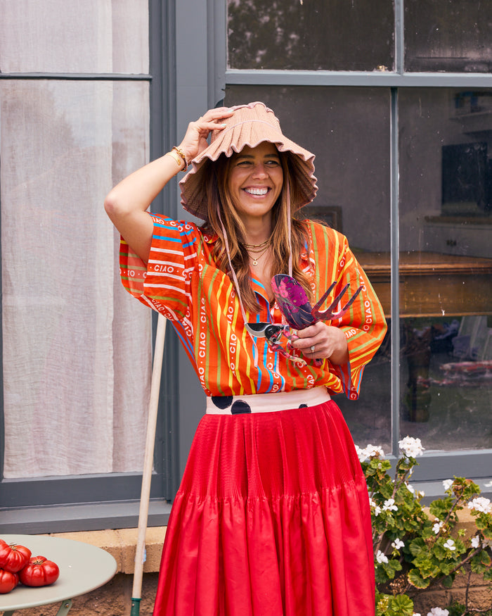 A woman stands outdoors in front of a window wearing a bright orange and red patterned blouse, a red skirt, and a beige wide-brim hat, holding a glass of water.