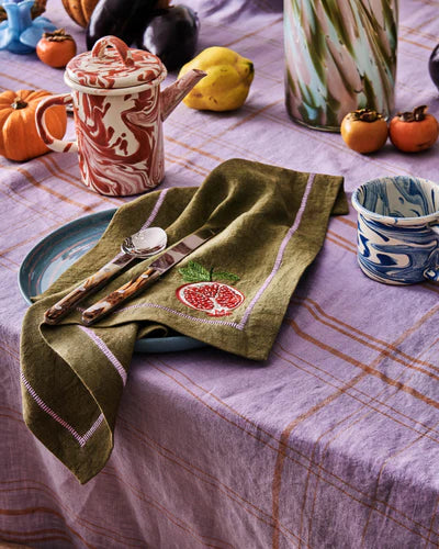 Green embroidered napkin with red pomegranate embroidery detail. Sat on top of blue plate and lilac tablecloth.