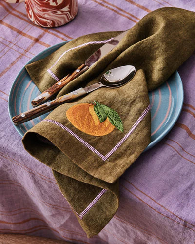 Embroidered khaki green napkin with orange and green leaf, sat on top of blue plate and lilac tablecloth. 