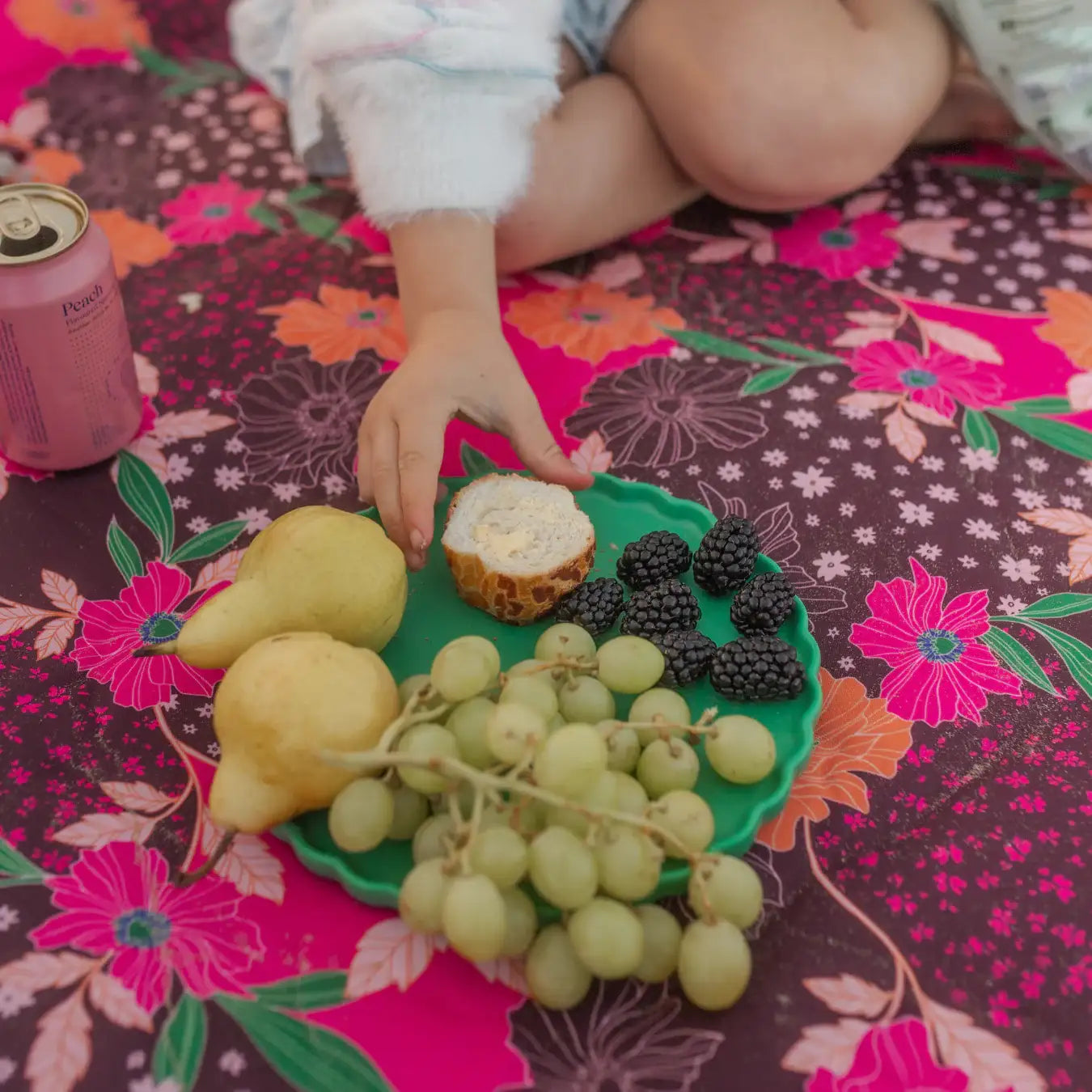 Child's hand reaching for a piece of bread on a plate of fruit on a floral tablecloth.