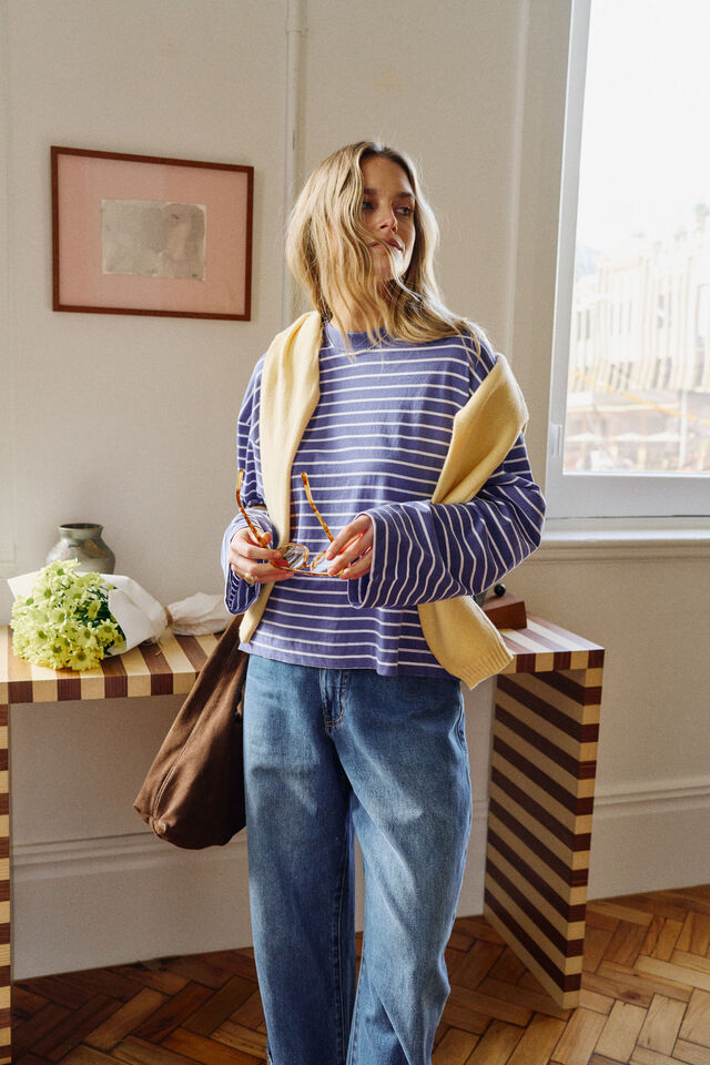A woman stands indoors wearing a blue and white striped long-sleeve top, blue jeans, and a beige cardigan draped over her shoulders, holding a brown tote bag.