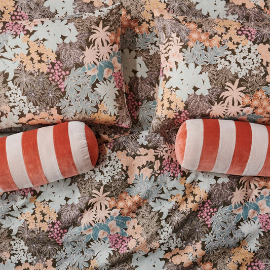 A fitted sheet with a floral pattern, predominantly in beige, brown, and multicolor tones, displayed on a bed with two cylindrical pillows with a striped pattern.