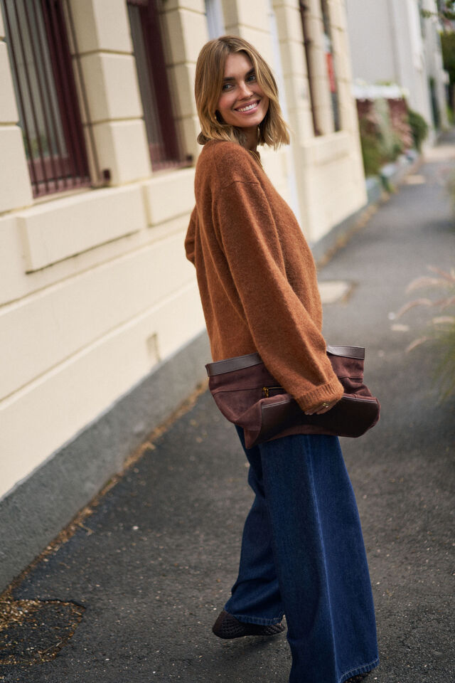 Woman in a brown sweater and blue jeans standing on a street.
