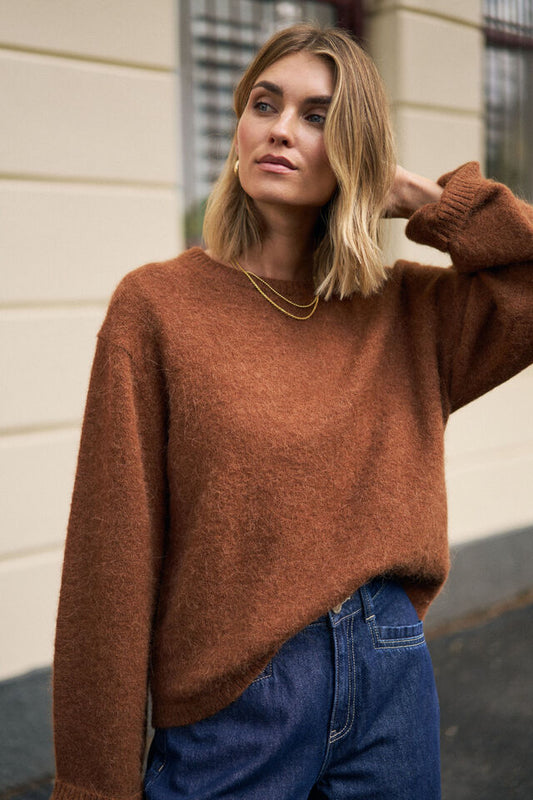 Woman wearing a brown sweater and blue jeans standing against a light-colored wall.