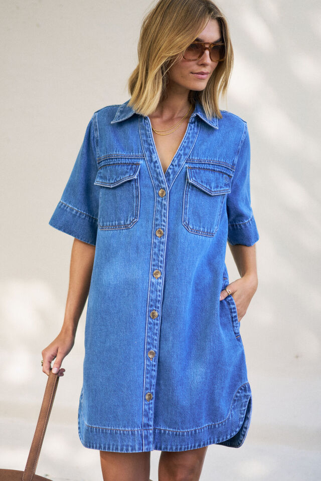 A woman wearing a blue denim shirt dress with short sleeves, a button-down front, and chest flap pockets, standing against a light neutral wall.