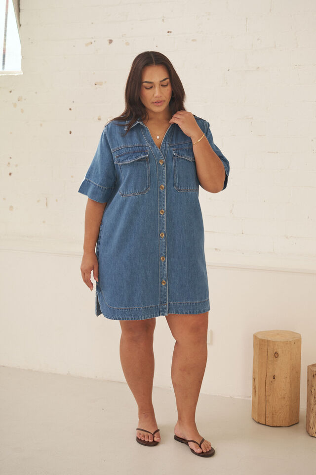A woman modeling a short-sleeve denim shirt dress with button-front closure and chest flap pockets, standing in a bright indoor room with a white brick wall and wooden stool props.