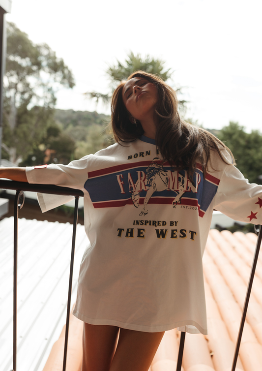 Person wearing a 'Born in the AUS, Inspired By the West' t-shirt on a balcony with trees in the background.