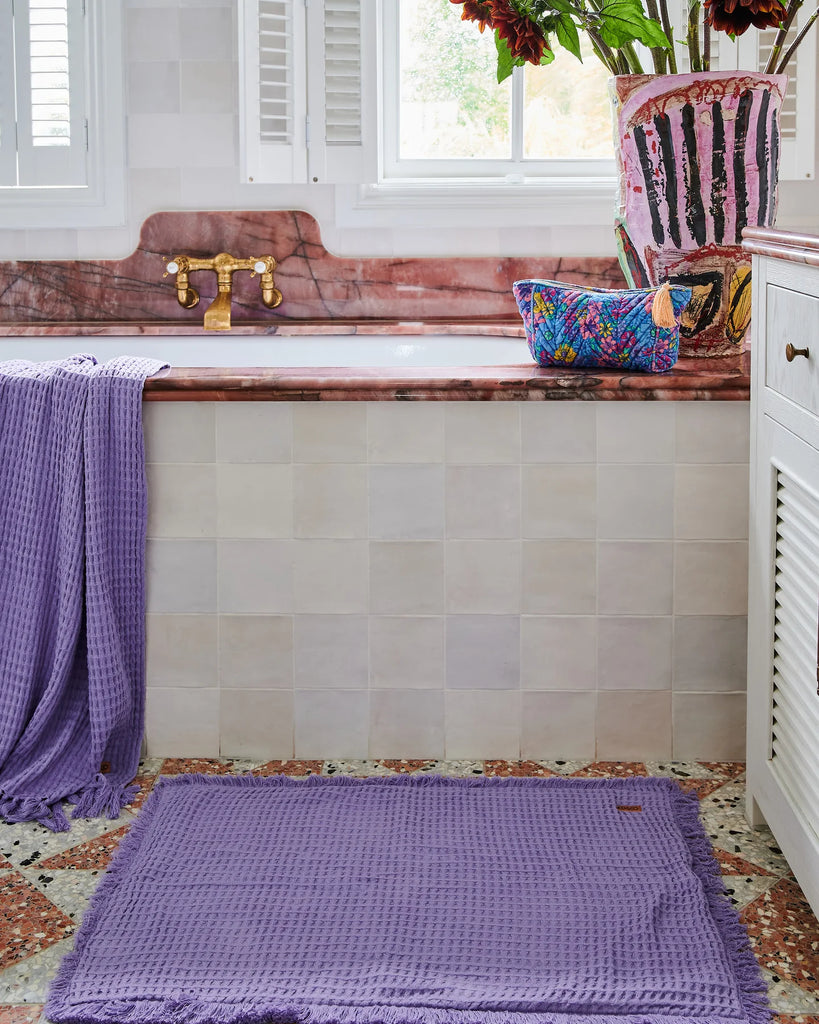 Toiletry bag sits on side of bath tub, next to vase and purple towel and bath mat 