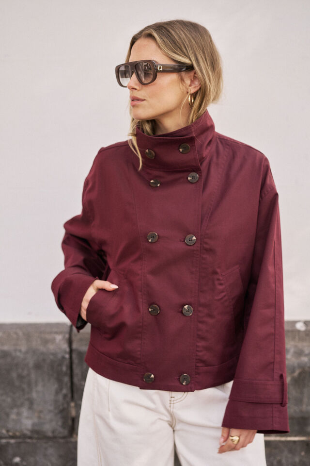 A woman wearing a cropped double-breasted jacket in deep burgundy with dark buttons, styled with white pants and sunglasses, standing against a neutral gray wall.