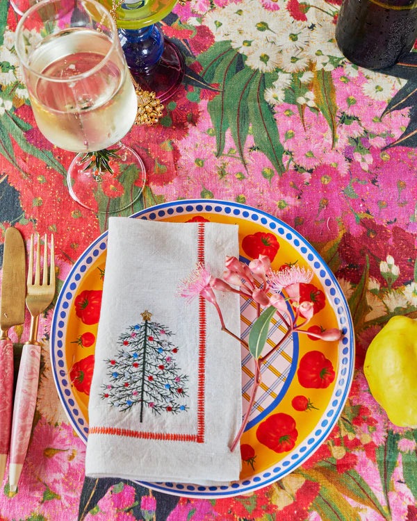 Close up of tablescape featuring bush blossom floral tablecloth in rich colours including green, pink, red and green. Also featured in a blossom sprig lying on top of plate, napkin, cutlery sits next to plate along with wine glass. 
