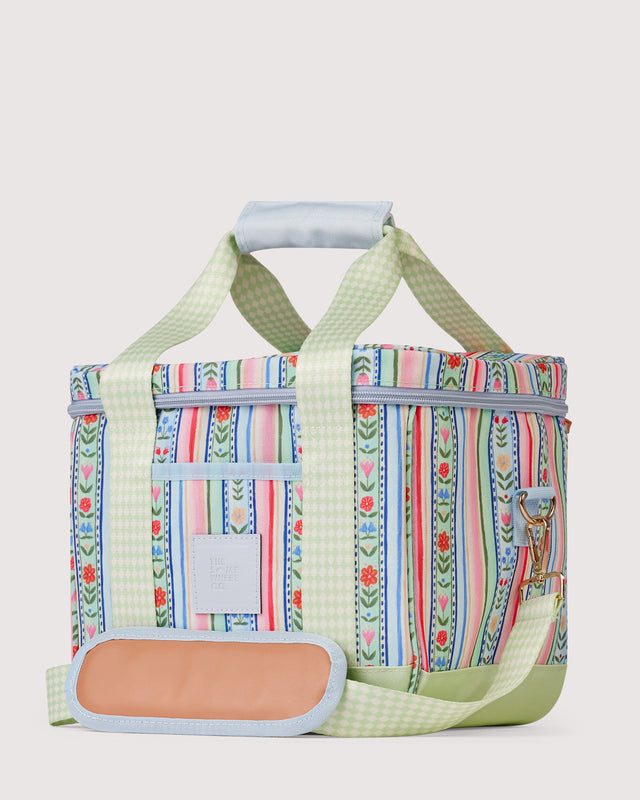 A patterned soft-sided cooler bag with light green gingham straps and trim, featuring vertical floral and striped prints in pink, blue, and green, with a peach-colored insulated pocket on the front.