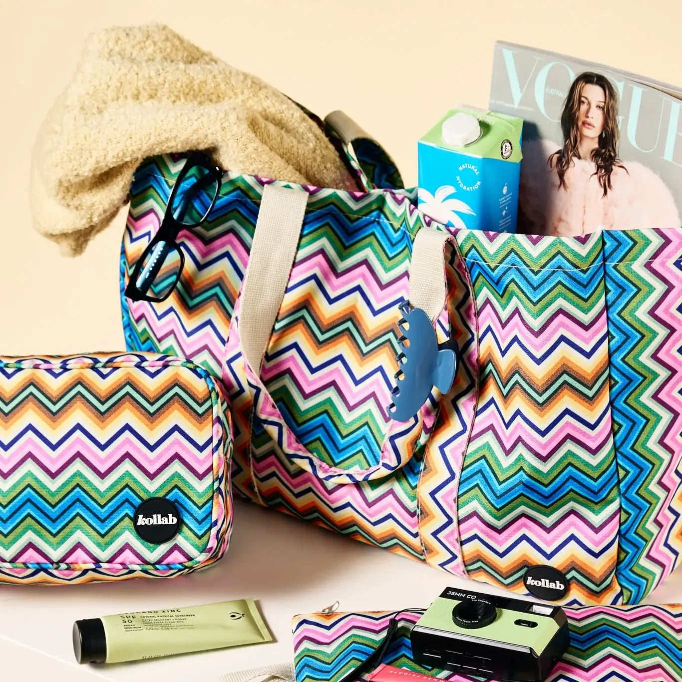 A colorful zigzag-patterned tote bag set with matching pouches, styled with travel accessories on a warm beige background. The set includes a large tote bag, a smaller matching pouch, and a zippered pocket, all featuring the 'kollab' brand logo.