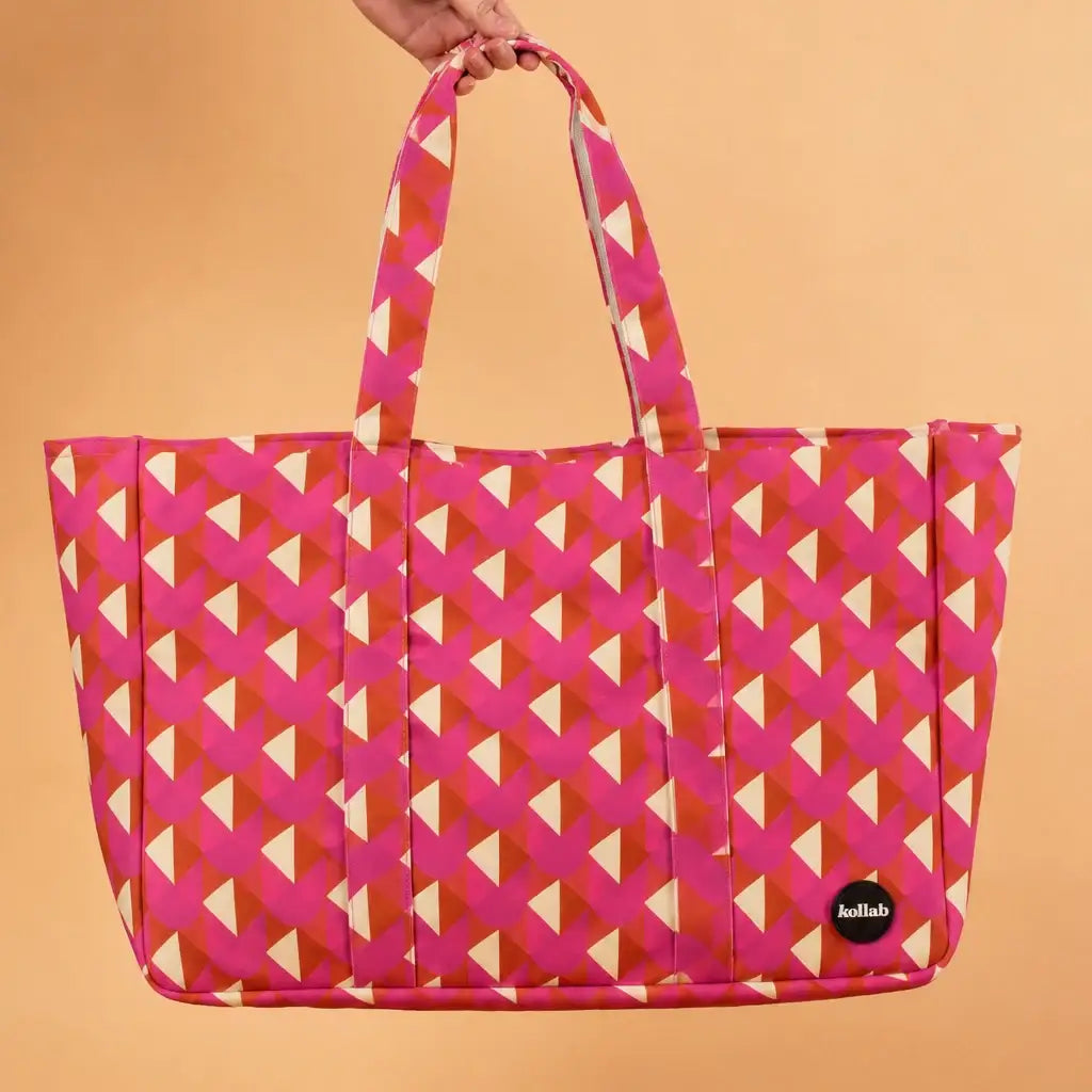 A pink tote bag with a geometric pattern of pink, orange, and cream triangles, held by a hand against a solid peach background. A small black circular 'ballab' logo patch is on the lower right corner of the bag.