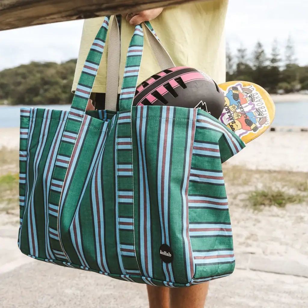 A green striped tote bag with white and blue vertical stripes, held by a person at a beach. The bag has a black circular patch with the brand name 'balab' on it, and is shown with a football and a skateboard tucked inside.