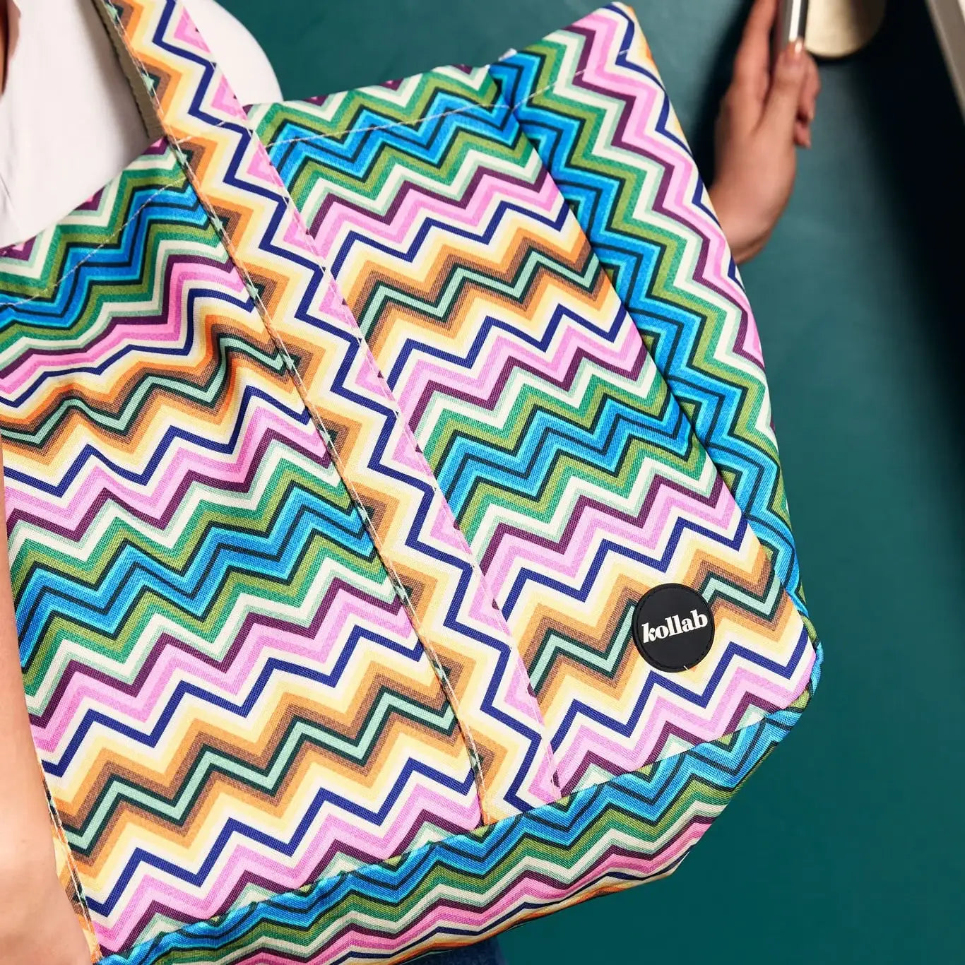 A close-up of a colorful zigzag-patterned tote bag with a black circular 'kollab' logo patch. The bag features a multicolor chevron design in pink, blue, green, yellow, orange, and purple.