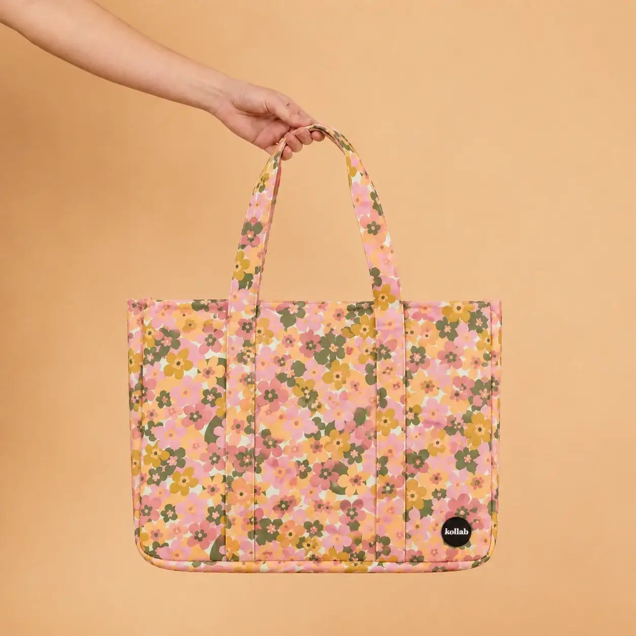 A person's hand holds up a pink floral tote bag with yellow, green, and white flowers against a solid peach background. A small black circular 'hololive' brand tag is visible on the bottom right corner of the bag.