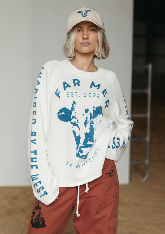 A model wearing a white crewneck sweatshirt with blue graphic prints and text, paired with rust-colored cargo pants and a beige baseball cap, standing in an indoor studio setting.