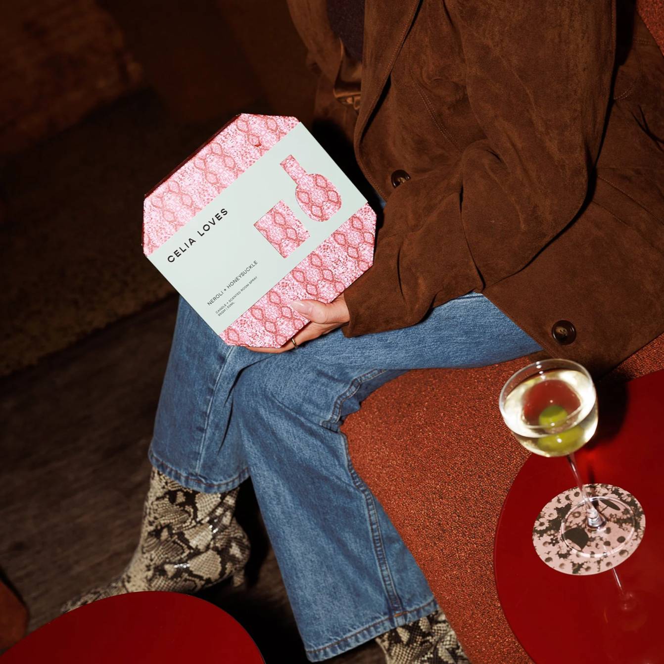 Person holding a pink and white patterned box with 'Celia Loves' branding, sitting on a red couch with a drink.