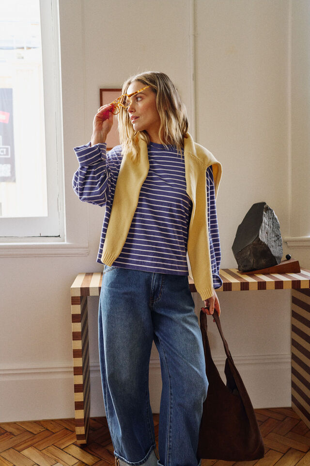 A woman wearing a navy and white striped long-sleeve top, a yellow sleeveless vest, and blue jeans, holding a brown tote bag in a bright indoor room.