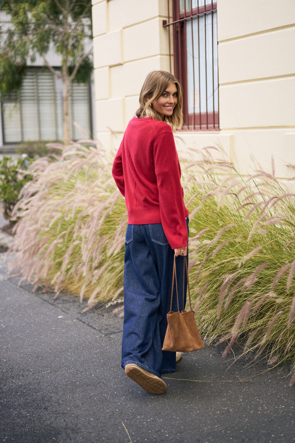 Woman in red sweater and blue pants walking on a street with decorative grasses.