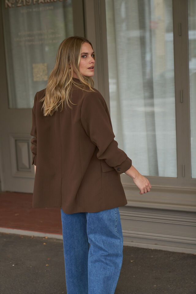 Woman wearing a brown jacket and blue jeans walking past a building entrance.