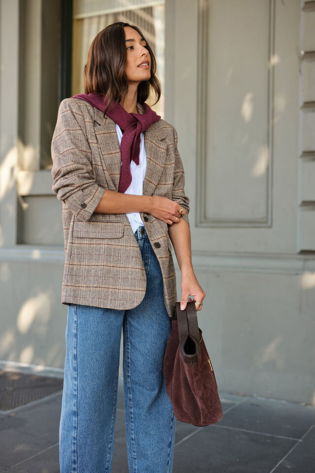 Woman wearing a plaid blazer, scarf, and jeans with a handbag, standing outdoors.