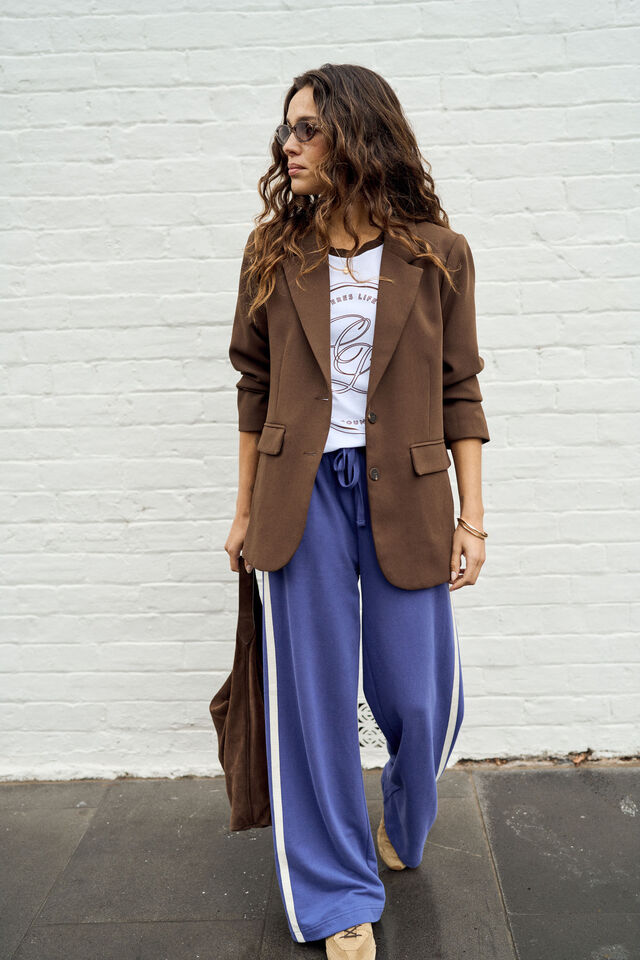 A woman stands against a white painted brick wall wearing a brown blazer over a white graphic T-shirt and blue wide-leg sweatpants with white side stripes. She is holding a brown bag.