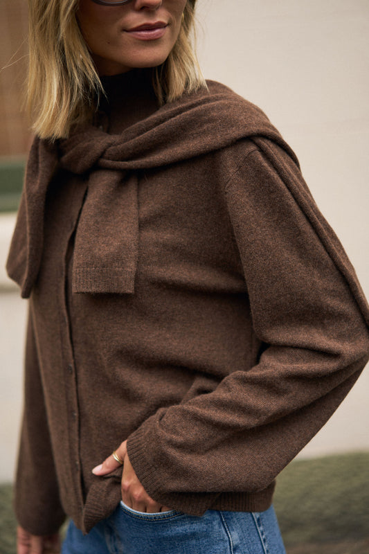 Brown sweater with another sweater across shoulders knotted at front worn by a woman with blonde short hair.
