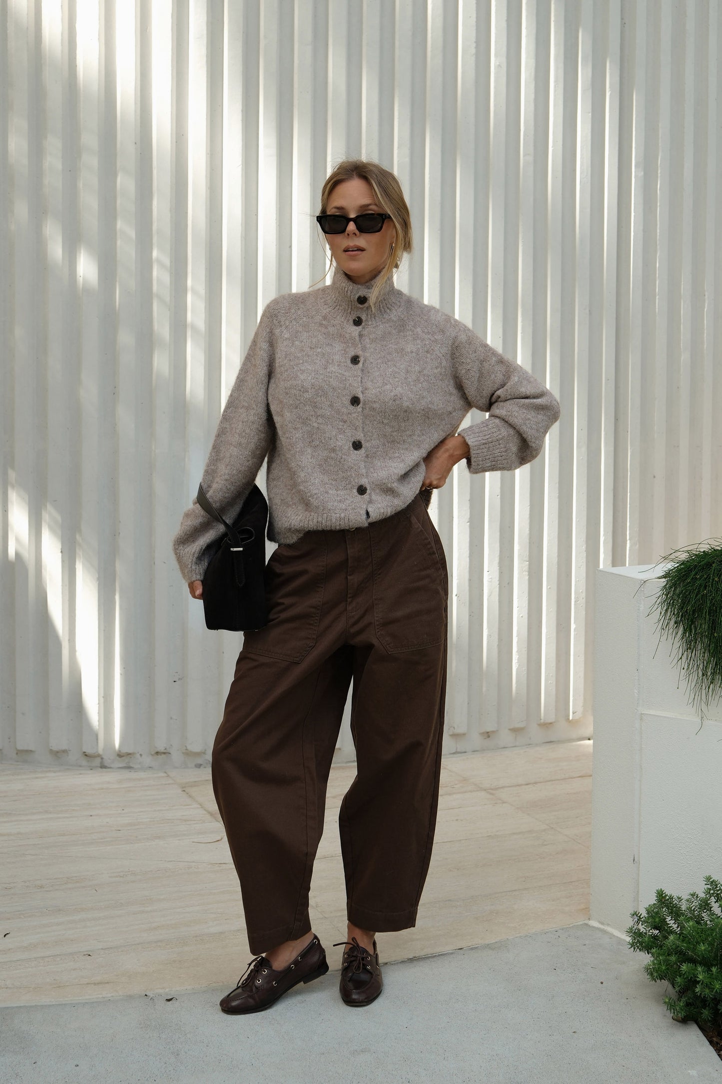 Woman wearing a beige sweater and brown pants standing against a white wall.