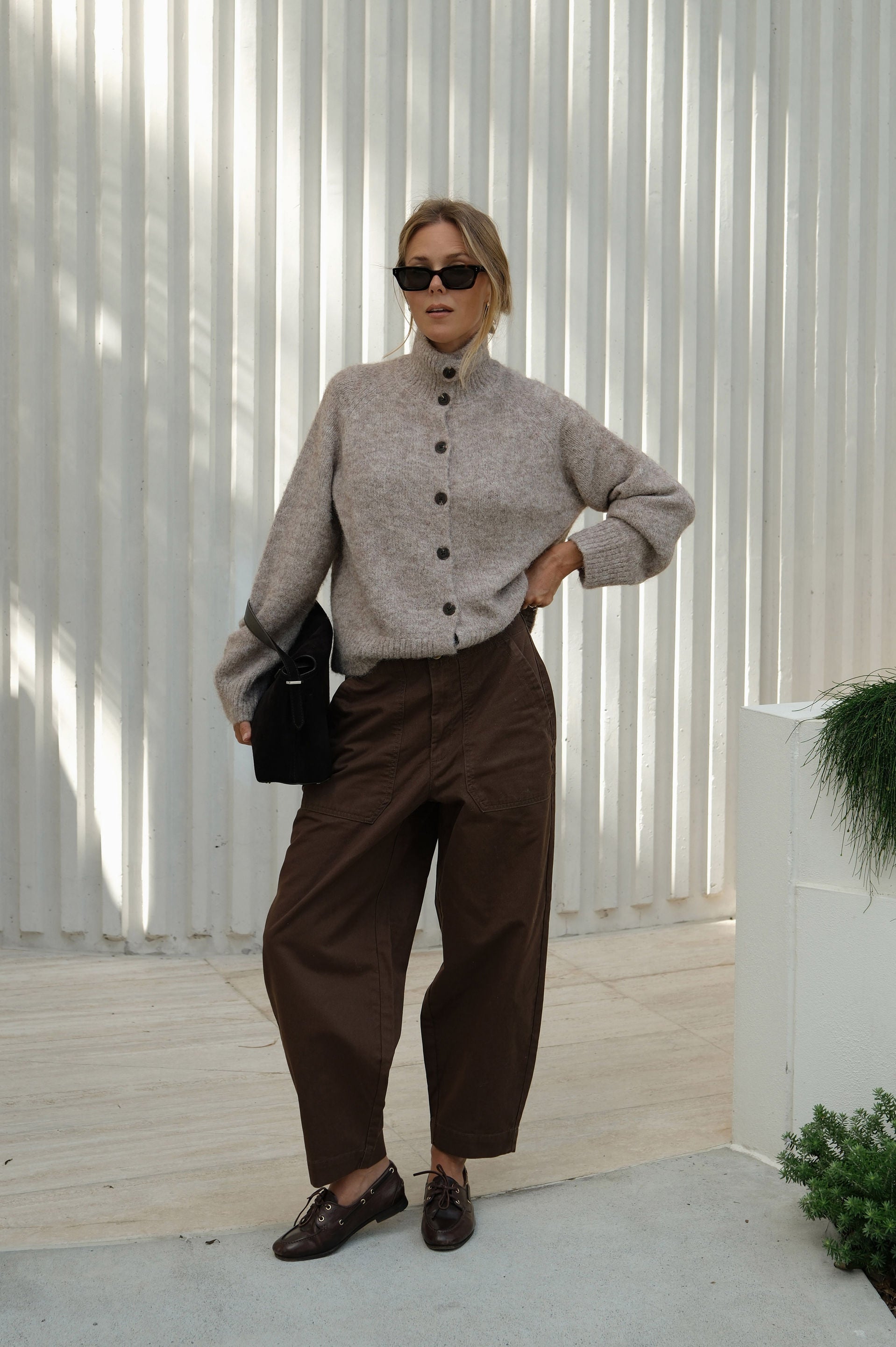 Woman wearing a beige sweater and brown pants standing against a white wall.