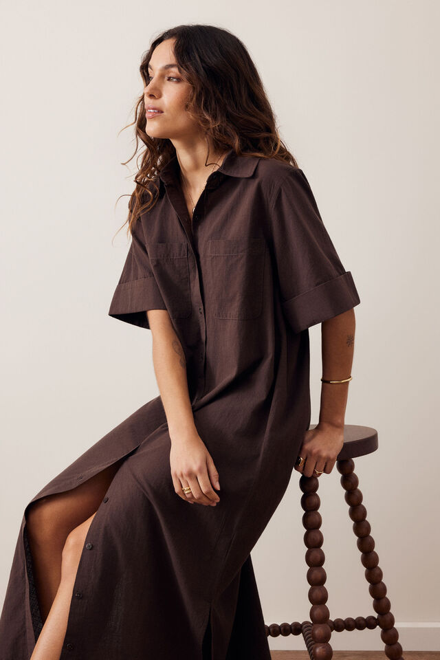 A woman wearing a dark brown short-sleeve collared dress sits on a wooden stool against a plain light beige wall.