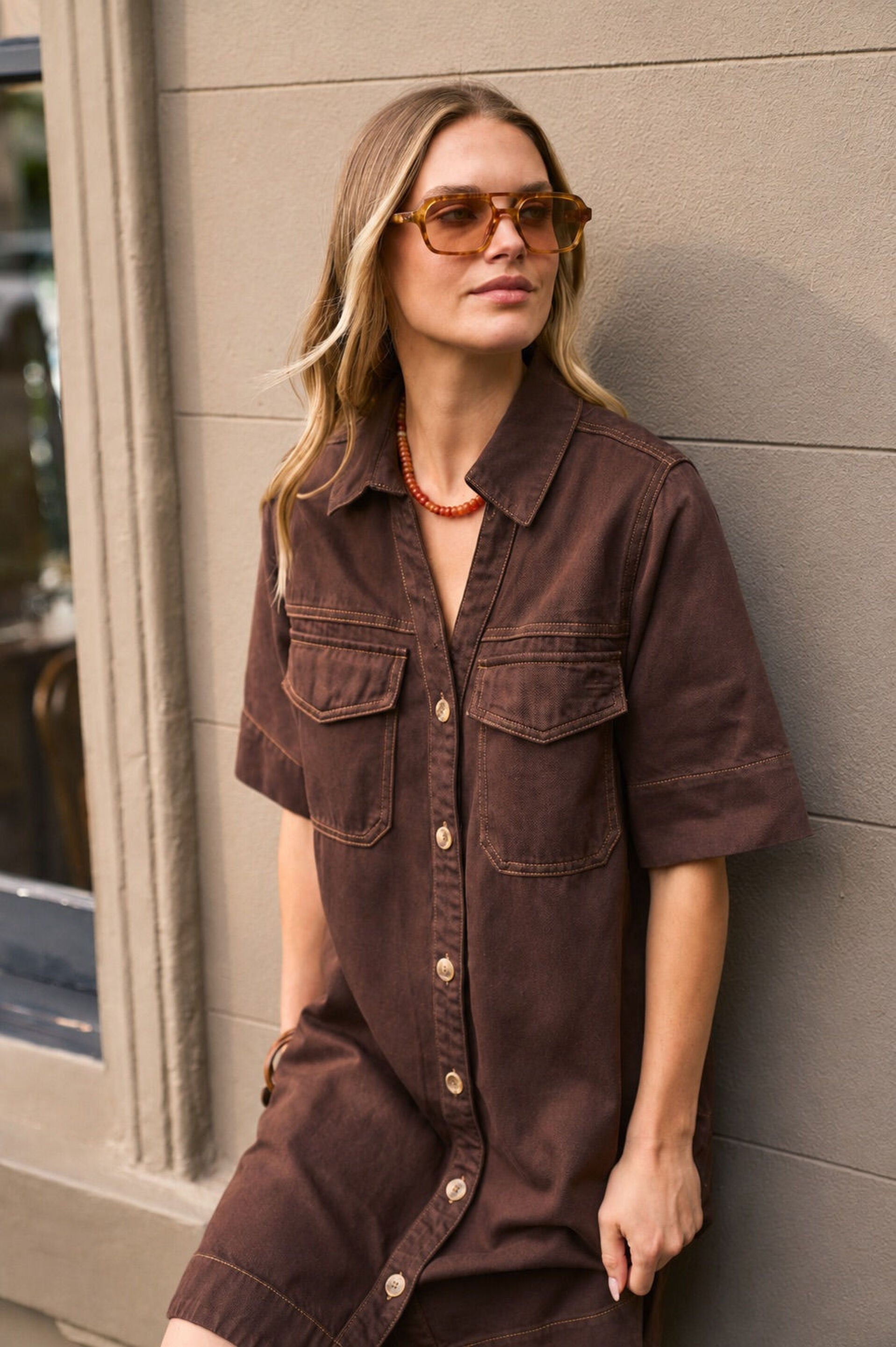 A woman wearing a dark brown short-sleeve button-front shirt dress leans against a gray paneled wall. She is wearing brown sunglasses and a red-orange beaded necklace.