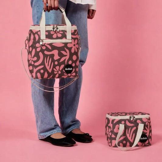 A person wearing blue jeans and black flats holds a pink and dark brown floral patterned tote bag with cream handles against a solid pink background. A matching zippered pouch sits on the floor beside them.
