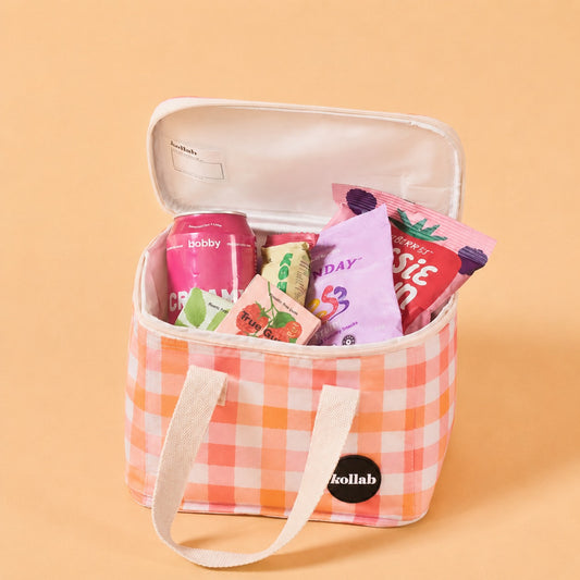 A pink and white gingham lunch bag with white canvas handles, shown open and filled with snacks and a can of soda. The bag has a black circular kollab logo patch on the front, set against a solid peach background.