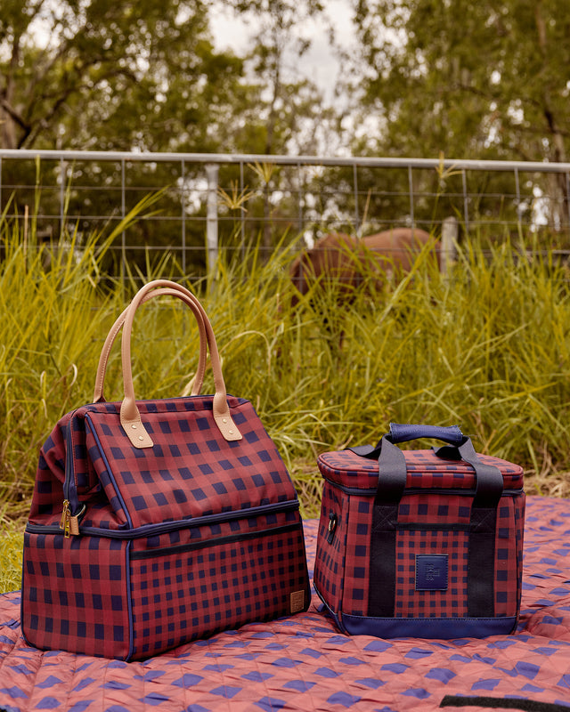 Two red and navy blue buffalo plaid insulated bags, one a tote bag with tan leather handles and one a lunch box with dark blue handles, sitting on a matching plaid blanket in an outdoor grassy field.