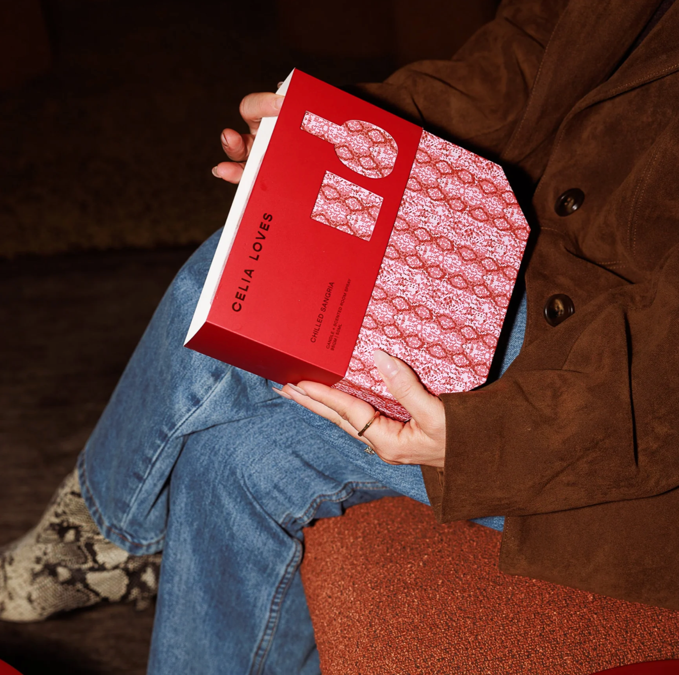 Person holding a red box with a patterned design, wearing a brown coat and blue jeans.