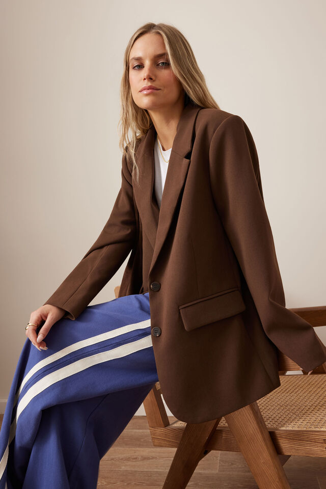 Woman wearing a brown suit with a blue skirt sitting on a wooden stool.