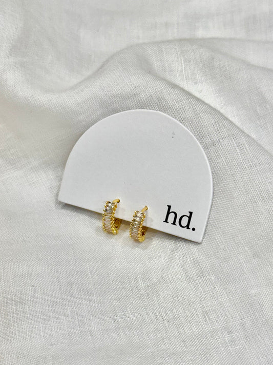 Gold earrings on a white card with 'hd.' text on a light gray fabric background
