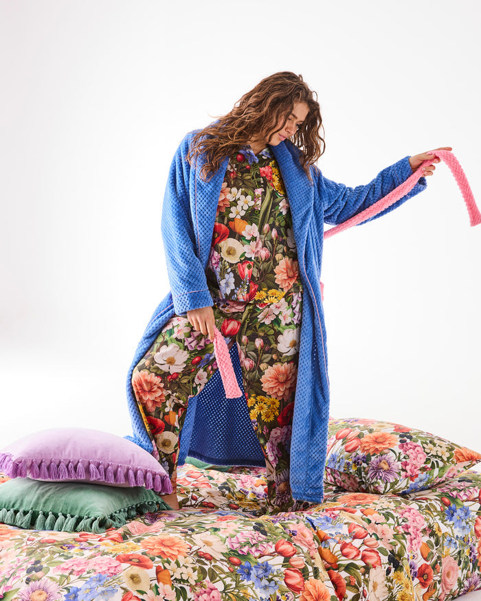 Woman wearing a blue robe over a floral pj set, standing on a bed with floral bedding.