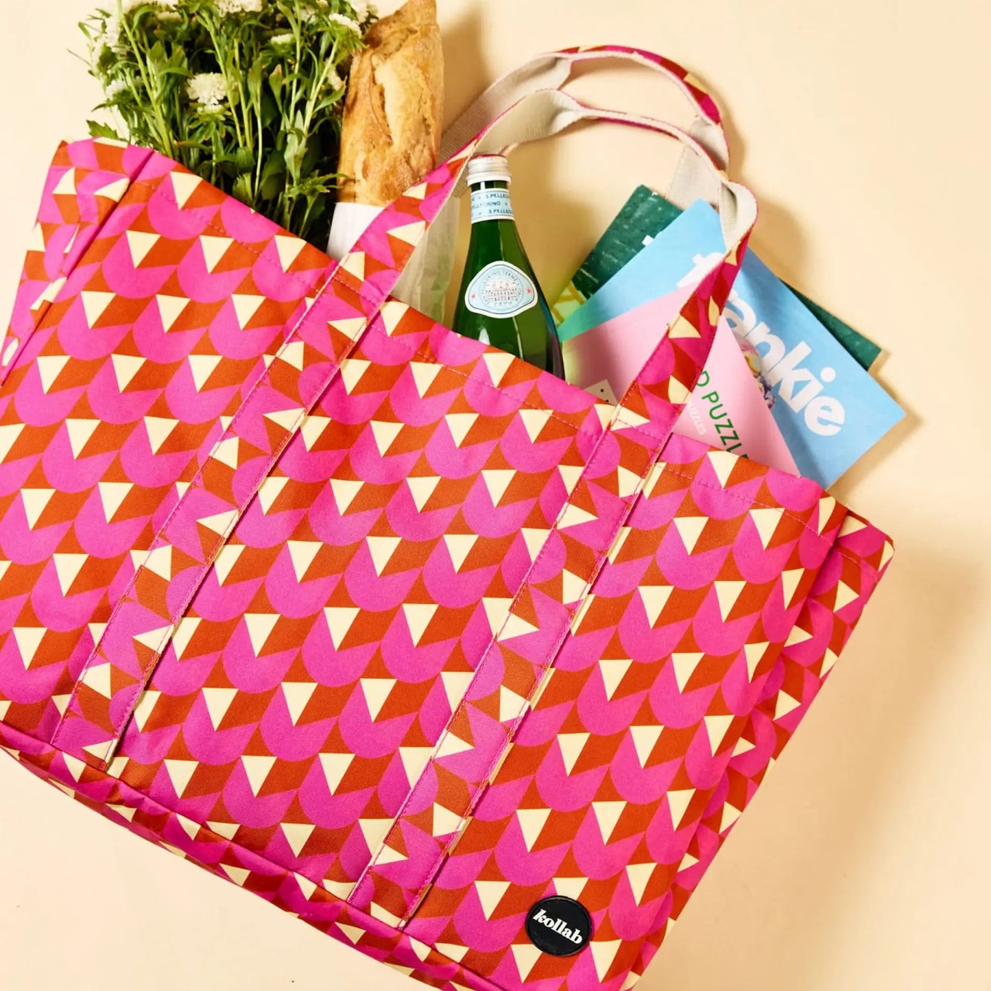A pink reusable tote bag with a geometric chevron pattern, shown filled with groceries including a green bottle, bread, and a puzzle book on a light beige background.