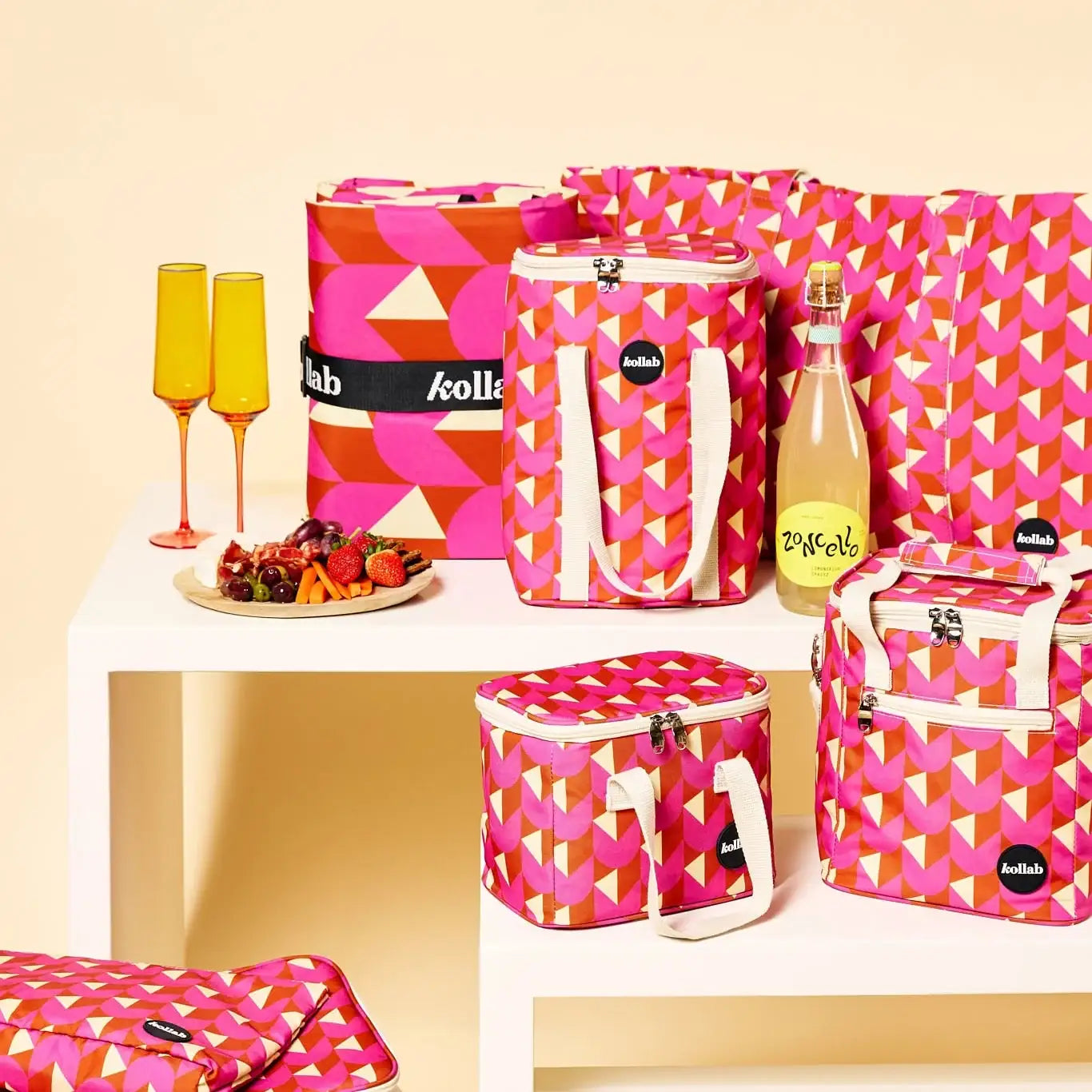 A coordinated set of pink geometric patterned picnic bags and insulated coolers with white handles, arranged on white shelves beside two orange-tinted champagne flutes and a bottle of sparkling wine. The bags have small black oval labels reading 'lab kollo'.