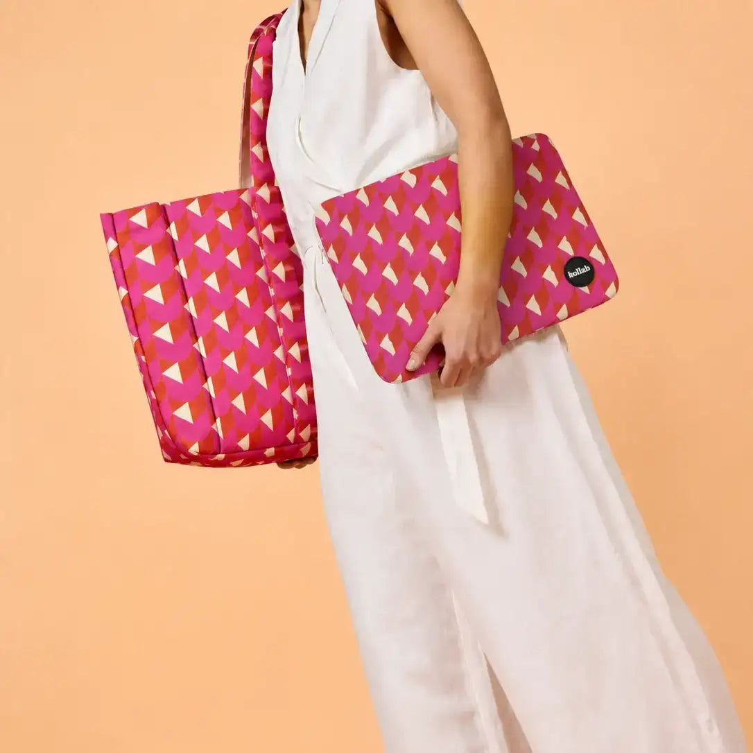 A woman in a white sleeveless dress holds a pink geometric patterned laptop sleeve and a matching tote bag against a solid peach background.