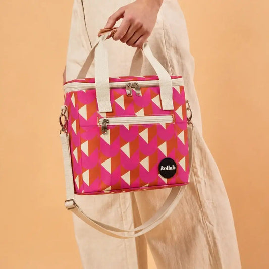 A person in beige clothing holds a structured cooler bag with a pink, orange, and white geometric pattern. The bag has cream-colored handles and a shoulder strap, silver zippers, and a black circular 'kallab' logo patch on the lower right front.