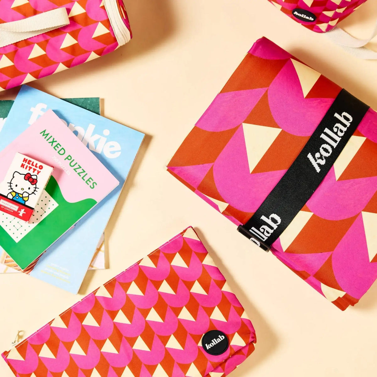 A styled flat-lay of reusable fabric pouches with a pink, orange, and cream geometric pattern, alongside a Hello Kitty puzzle book. The pouches feature black elastic bands with the white 'kollab' brand name.