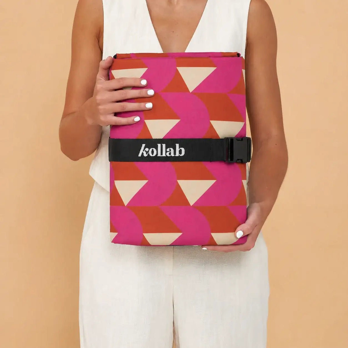 A model in a white sleeveless jumpsuit holds a folded tablet case featuring a vibrant geometric pattern of pink, orange, and beige triangles. The case is secured with a black horizontal strap across the middle that has the brand name 'kollab' printed in white.