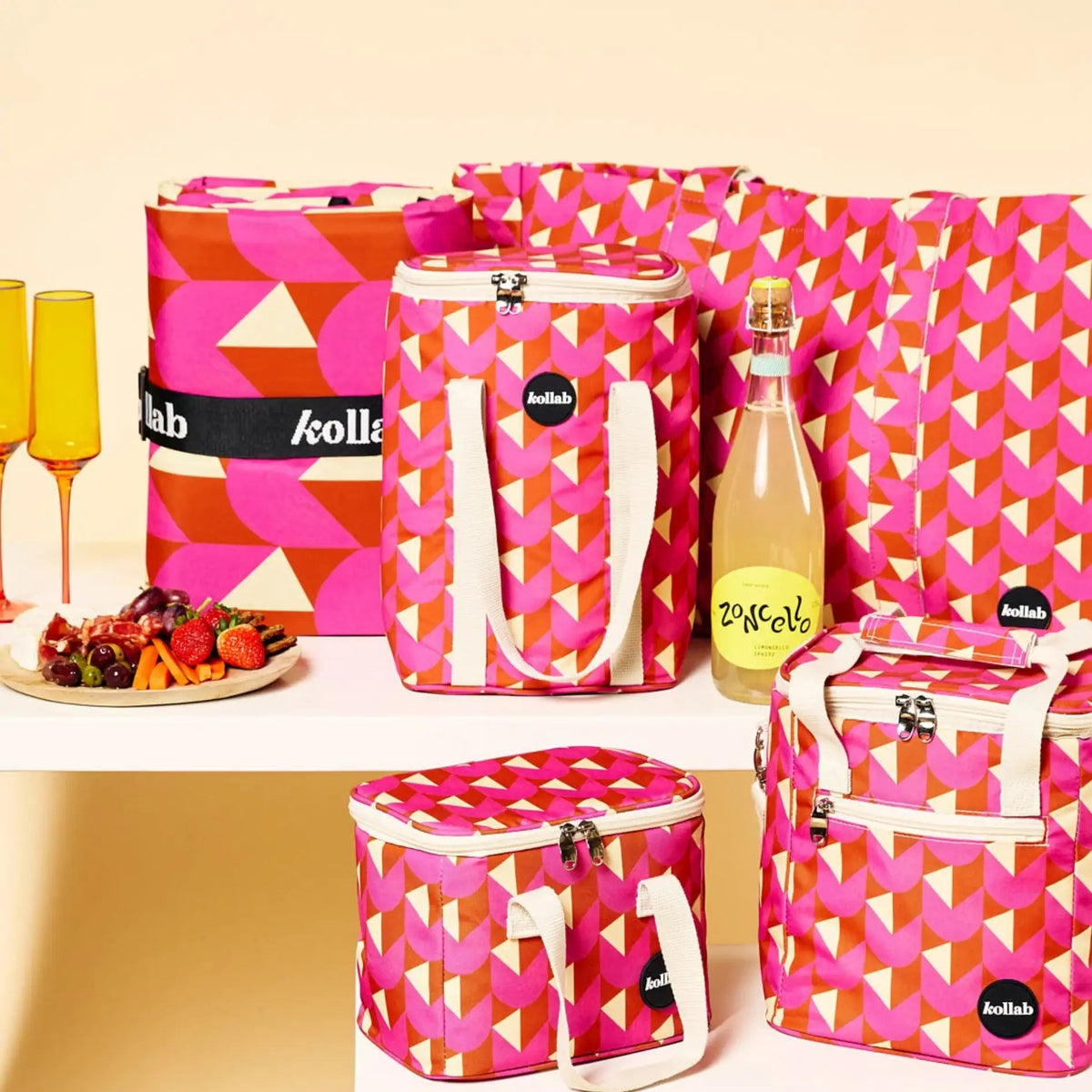 A set of Kollé fashion accessories in a matching pink, orange, and white geometric pattern, including insulated bags, a tote bag, a bottle, and a plate of fruit, arranged on a white shelf against a warm beige background.